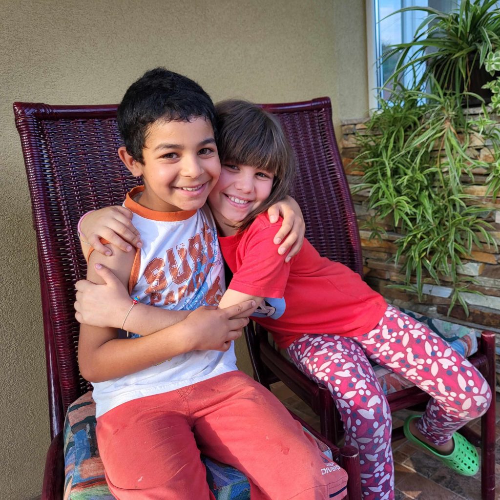 Ein Junge und ein Mädchen (Geschwister) im Grundschulalter sitzen nebeneinander, umarmen sich und strahlen in die Kamera