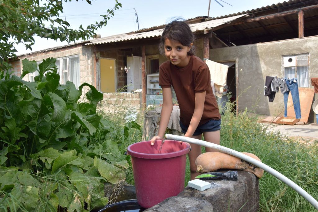 Ein Mädchen in braunem T-Shirt und kurzer Jeans steht vor einem maroden Haus und lässt Wasser aus einem Schlauch in einen Eimer laufen