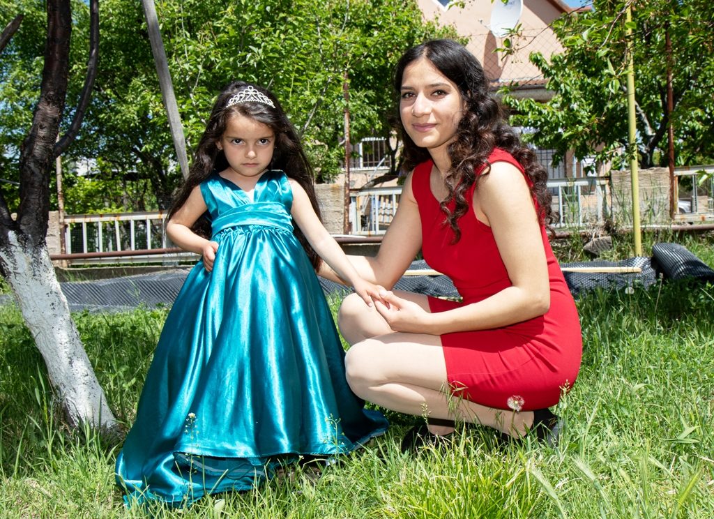 A young woman with long black hair and a red shift dress is crouching on the grass next to a little black-haired girl in a turquoise evening dress with a tiara on her head