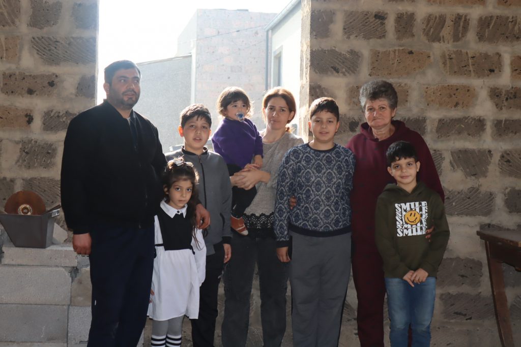 Drei Erwachsene und fünf Kinder stehen in einem Raum vor einer Fensteröffnung, in der jedoch das Fenster fehlt