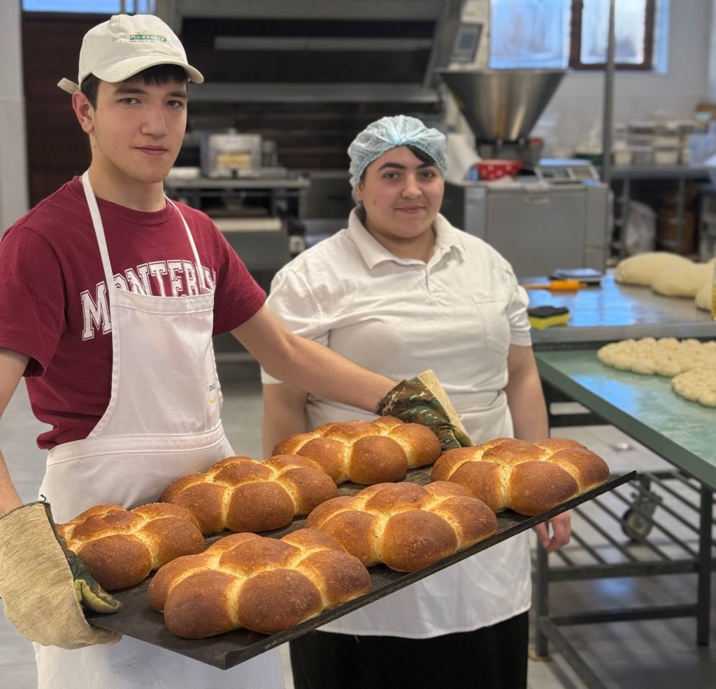 Ein junger Mann und eine Frau in Berufskleidung stehen nebeneinander in einer Bäckerei; der Mann hält ein Blech mit Broten