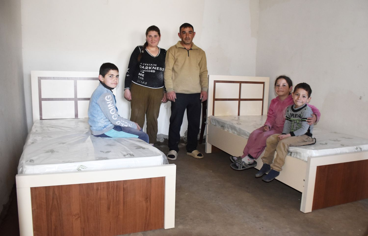 Eine Familie in einem Schlafzimmer; drei Kinder sitzen auf zwei Betten, die links und rechts an der Wand stehen, die Eltern stehen dazwischen an der Rückwand