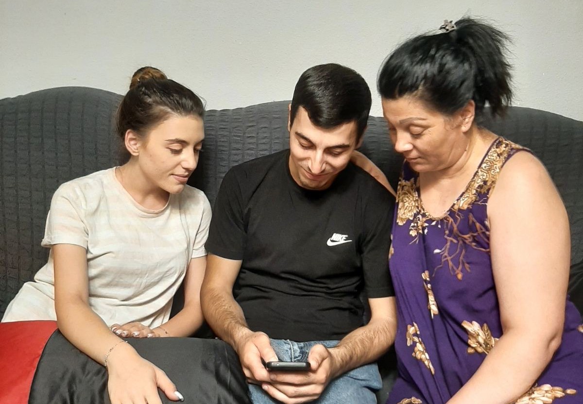 Eine Frau, ein junger Mann und eine junge Frau sitzen auf einem Sofa und schauen auf ein Smartphone.