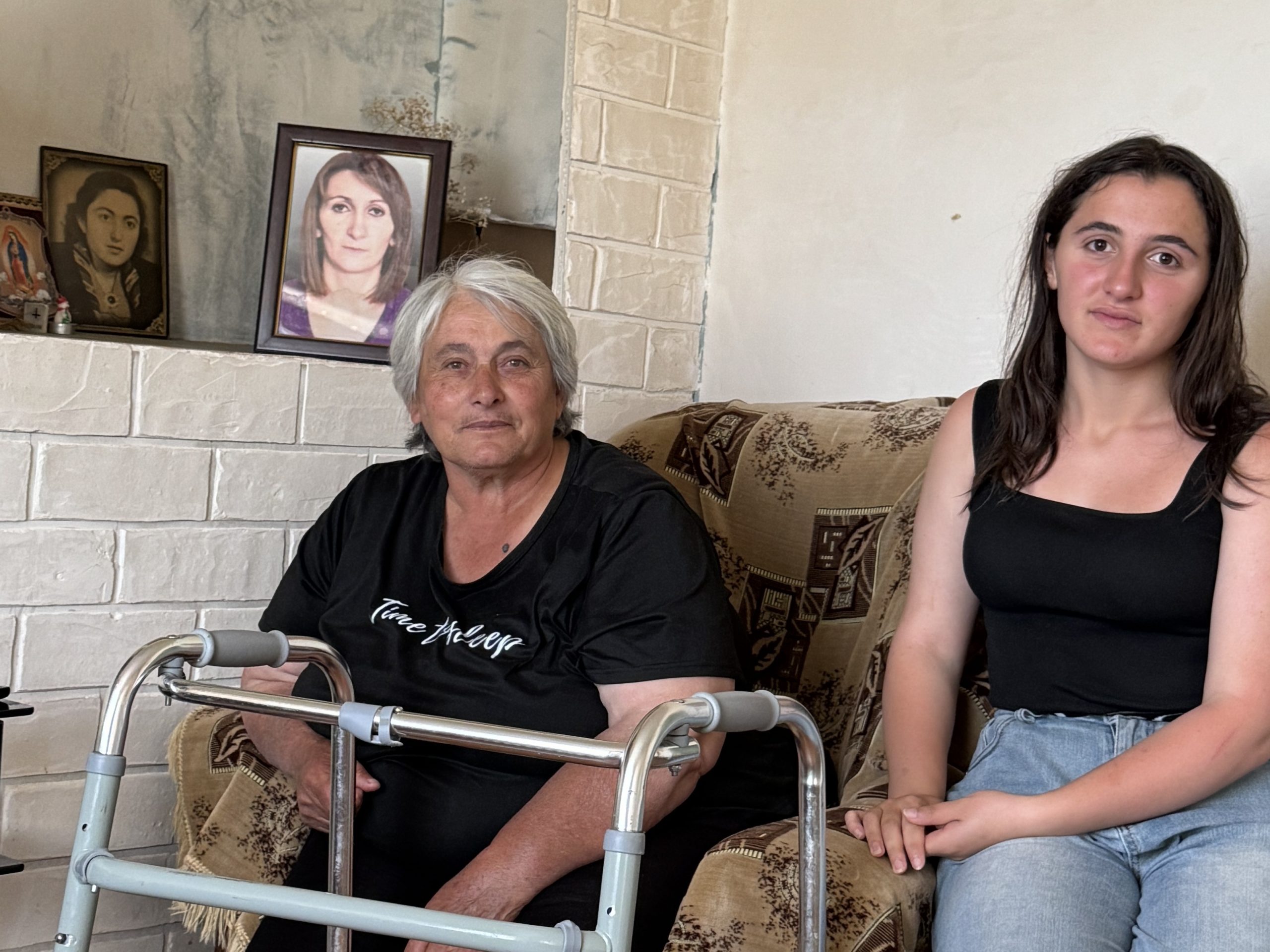 Eine ältere Frau und ein junges Mädchen sitzen auf einem Sofa. Die ältere Frau hat eine Gehhilfe. Im Hintergrund steht ein Foto einer jüngeren Frau.