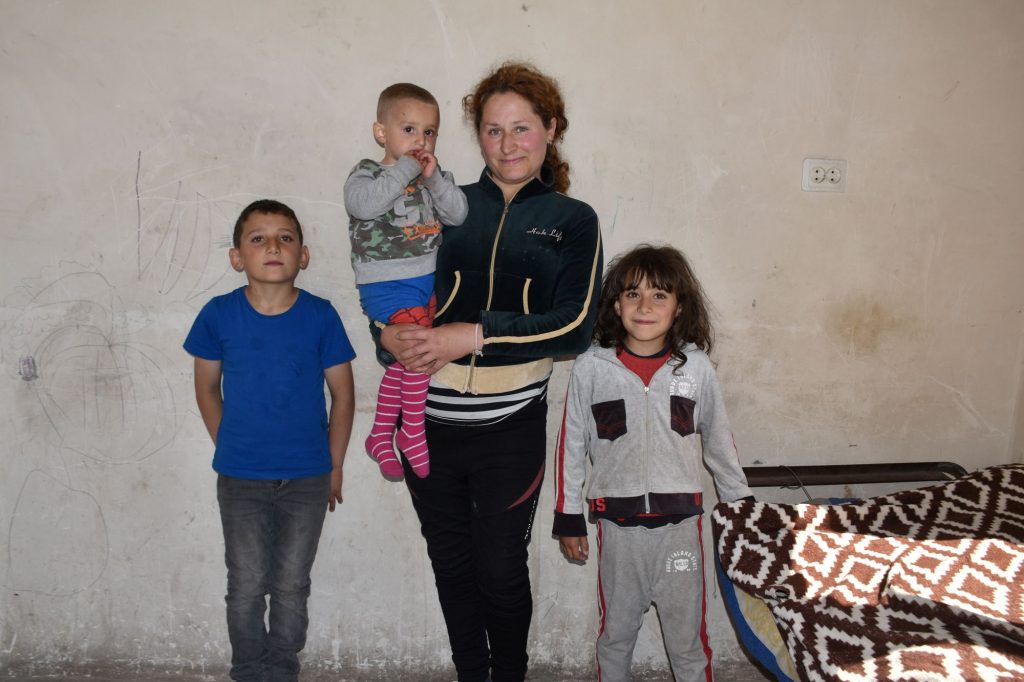 Eine Frau steht vor einer kahlen Wand, rechts von ihr ein Junge, links ein Mädchen. Auf dem Arm trägt sie einen kleinen Jungen.