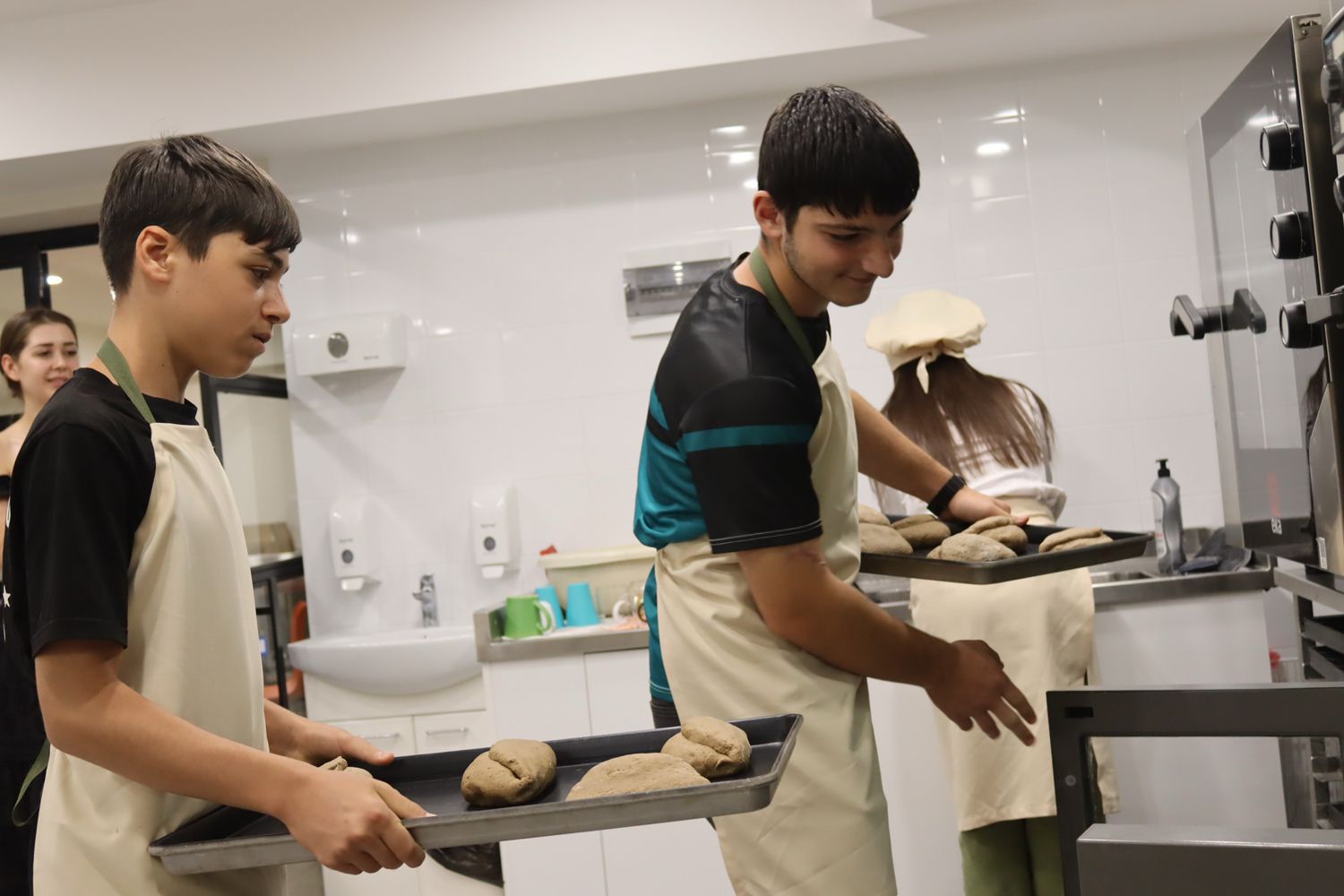 Zwei Jugendliche in der Bäckerausbildung; der Junge links trägt ein Blech mit Backwaren