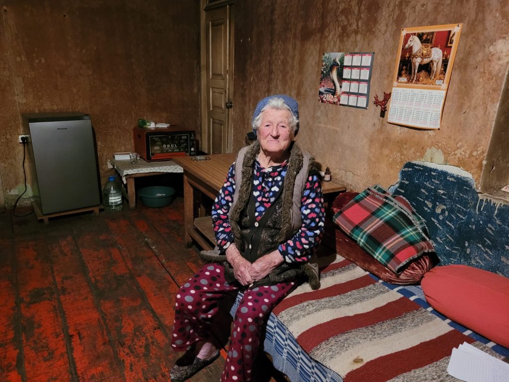 Eine ältere Frau sitzt auf ihrem Bett in einem Zimmer mit bräunlichen Wänden