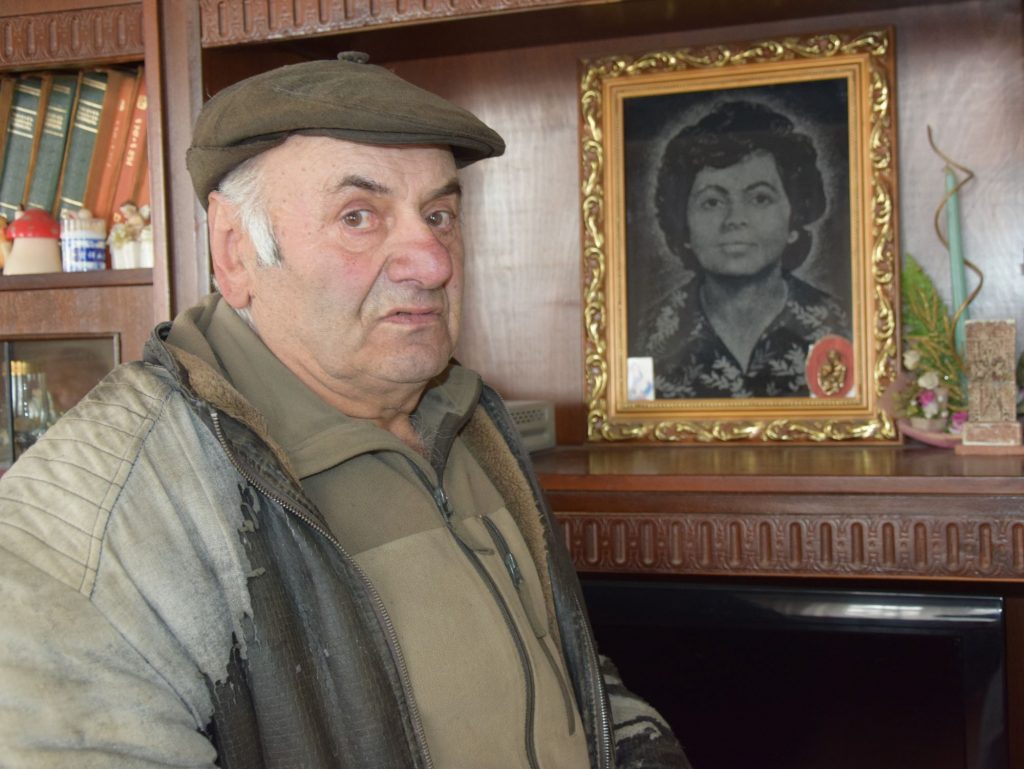 Ein älterer Mann steht neben dem Bild seiner früh verstorbenen Frau