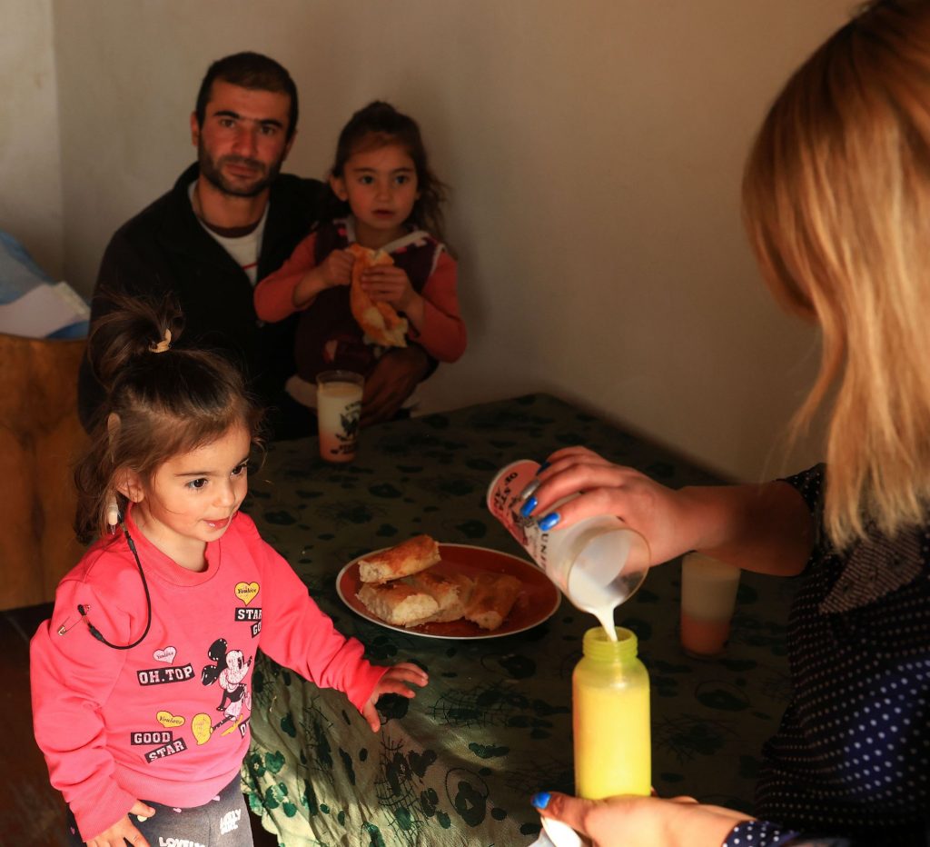 Eine Frau schüttet Milch in eine Trinkflasche, mit Spannung beobachtet von einem kleinen Mädchen. Im Hintergrund sitzt ein Mann mit einem kleinen Mädchen auf dem Schoß.