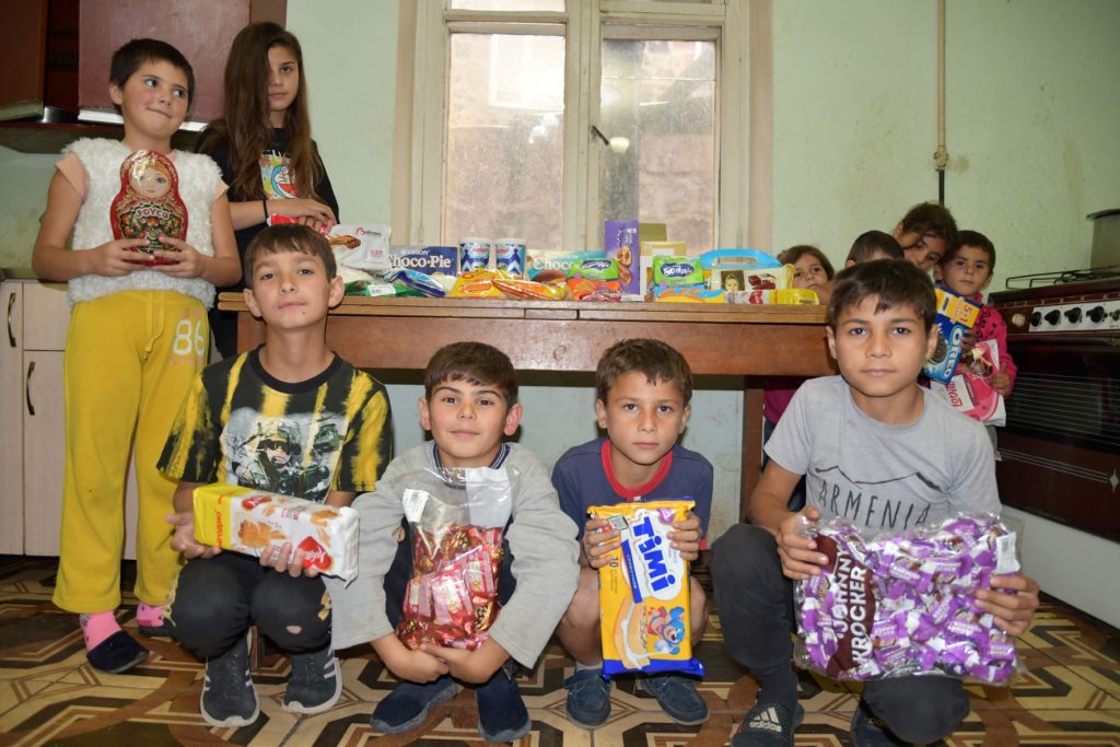 Ten children stand or squat around a table holding packages of groceries and candy. There are more packs on the table.