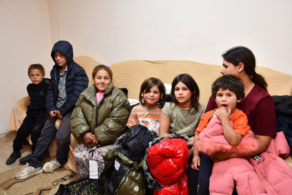 A mother and six children are sitting on a sofa. Some of the children are wearing winter coats.