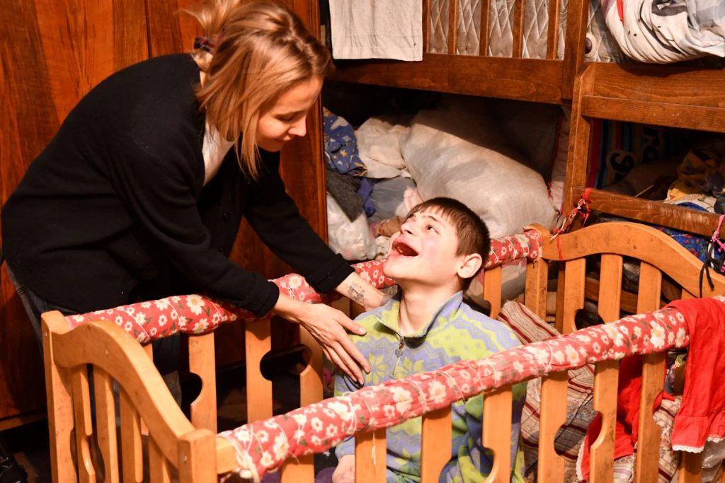 A teenage boy with a severe disability is sitting in a cot and smiles at a woman who bends down to him.