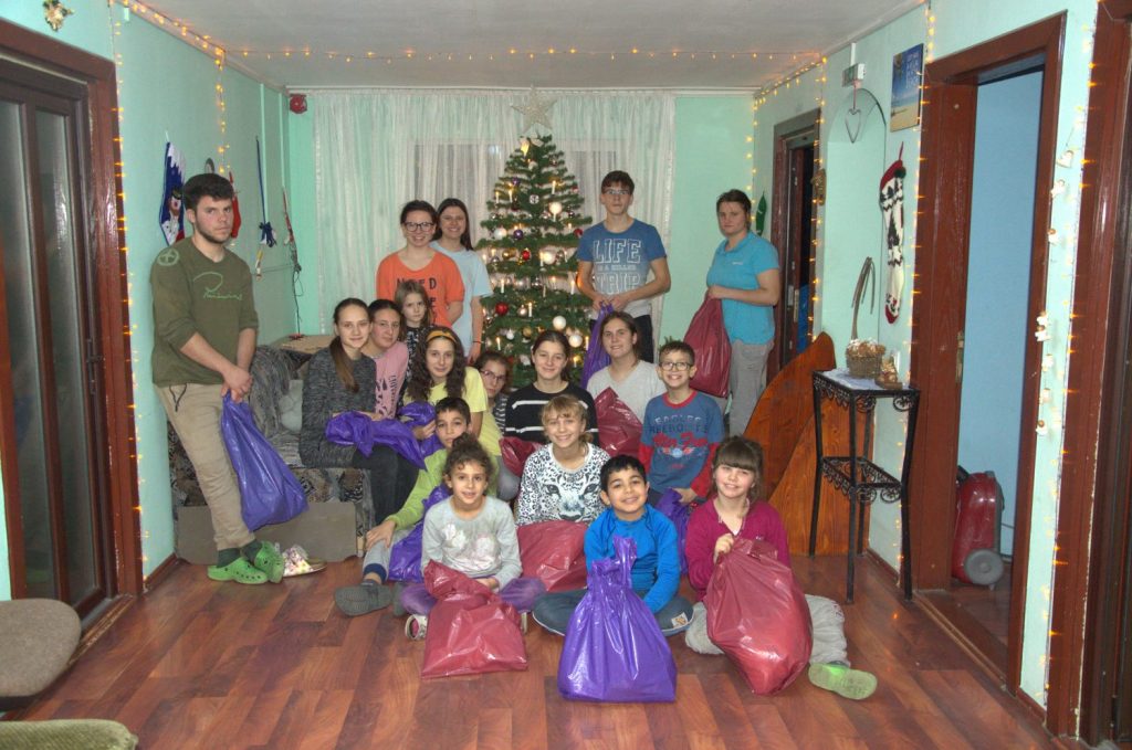 Several children and teenagers are sitting in a room around a Christmas tree and are holding large plastic bags in their hands.