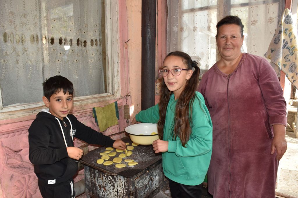 Eine Frau, ein Mädchen und ein Junge stehen an einem Ofen. Die Kinder legen Kartoffelscheiben auf die Oberfläche.