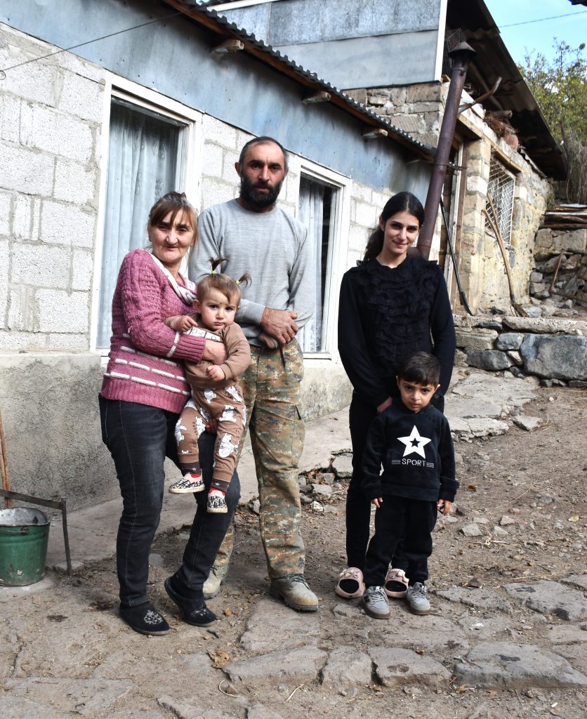 Ein Mann und eine Frau mittleren Alters sowie eine junge Frau stehen vor einem maroden Haus. Die ältere Frau trägt ein kleines Mädchen, ein kleiner Junge steht vor der jungen Frau.