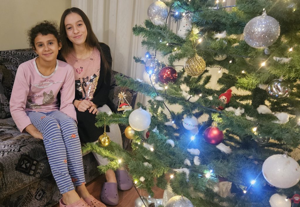 Zwei junge Mädchen sitzen neben einem Weihnachtsbaum auf dem Sofa und lächeln in die Kamera.